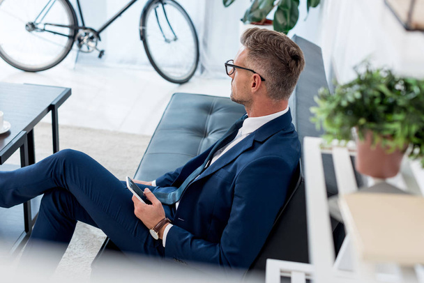 selective focus of businessman in glasses sitting on sofa and holding smartphone - Valokuva, kuva