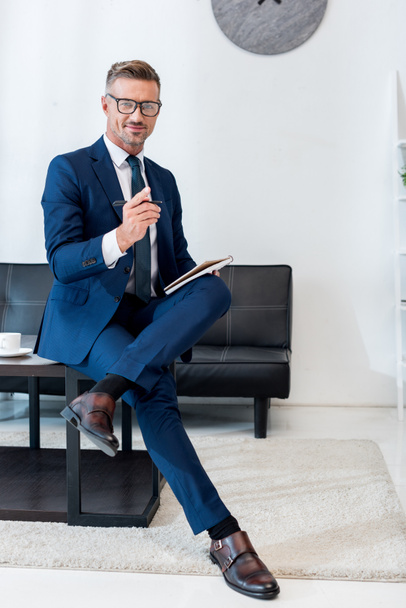 cheerful businessman in glasses holding notebook and pen while sitting on coffee table with crossed legs - Photo, Image