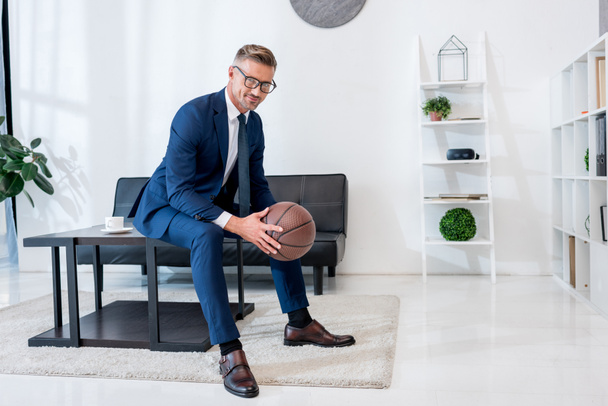 cheerful businessman in suit holding basketball in hands while sitting on coffee table - Photo, Image