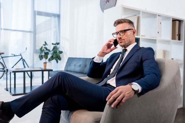 podnikatel v brýlích sedí v křesle a mluví na smartphone - Fotografie, Obrázek