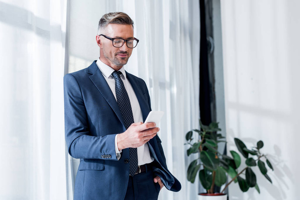 cheerful businessman in suit and glasses standing with hand in pocket and using smartphone - Foto, Imagen