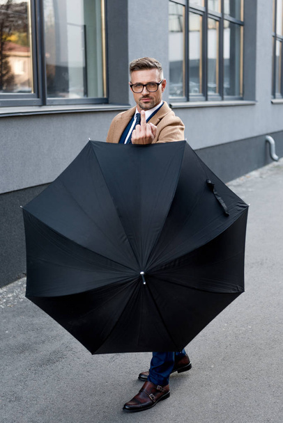 guapo hombre de negocios en gafas mostrando el dedo medio mientras sostiene el paraguas
  - Foto, Imagen