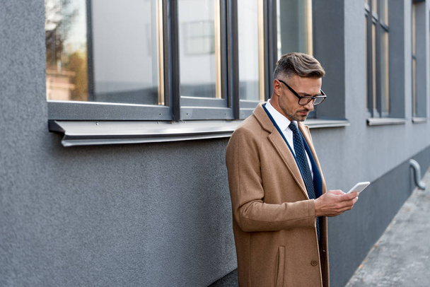 confident businessman in glasses looking at smartphone while standing in beige coat near building - Foto, Bild