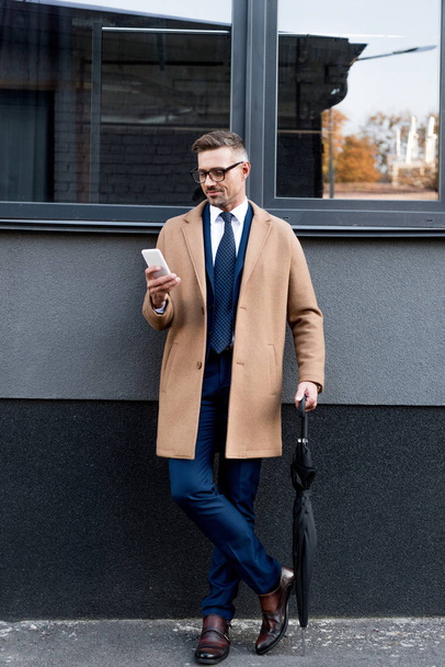 empresário alegre em óculos olhando para o smartphone enquanto de pé no casaco bege e segurando guarda-chuva
  - Foto, Imagem