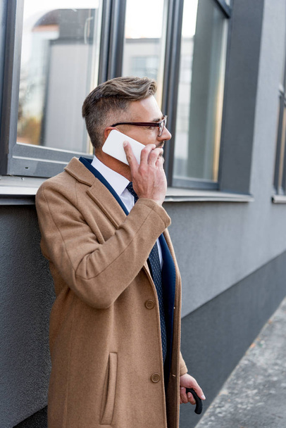bel homme d'affaires en lunettes parlant sur smartphone tout en restant debout en manteau beige
  - Photo, image