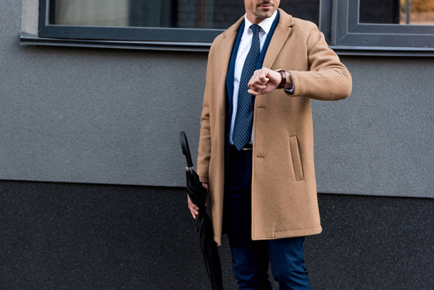 περικοπεί προβολή του επιχειρηματία, κοιτάζοντας το ρολόι και κρατώντας ομπρέλα  - Φωτογραφία, εικόνα