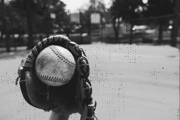 El viejo béisbol usado atrapado en un guante. Deporte mostrado en blanco y negro con campo de pelota en fondo y textura vintage
. - Foto, Imagen