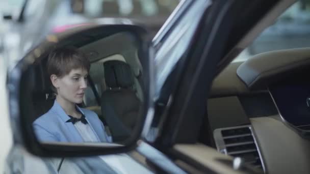 Beautiful young girl with a short haircut examines new auto while sitting in passenger compartment of the new car in car showroom. Successful pretty woman examines new auto. Car dealership. - Felvétel, videó