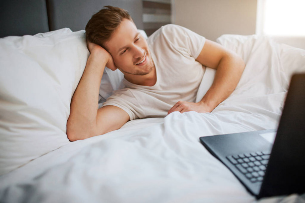 Young cheerful man lying in bed erly morning. He smile. Msle model look at laptop screen. Guy has rest. Daylight. - Foto, Imagem