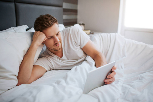 Relaxed peaceful young man lying in bed this morning. He hold white tablet. Guy look and it and smile. Man hold head on hand. Calm and peaceful. - Fotografie, Obrázek