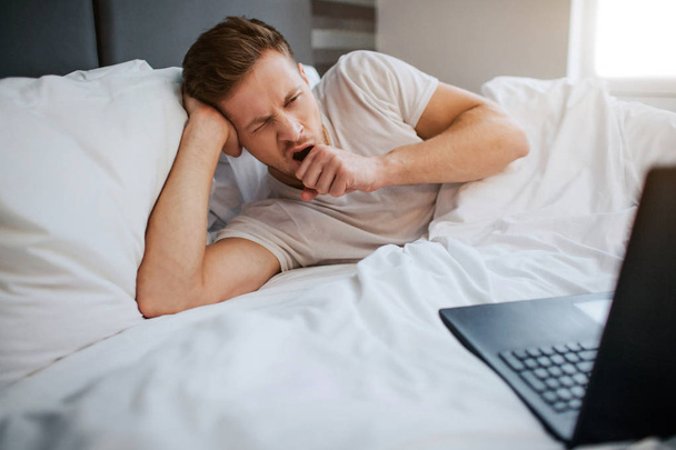 Young tired man lying in bed this morning. He yawn. Guy is sleepy. Young man look on laptop. - Photo, Image