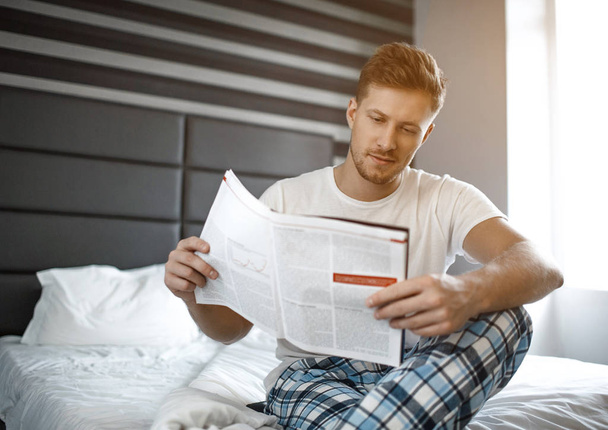 Young man on bed early morning. He sit and read journal. Small smile on face. Concentrated. - Foto, immagini