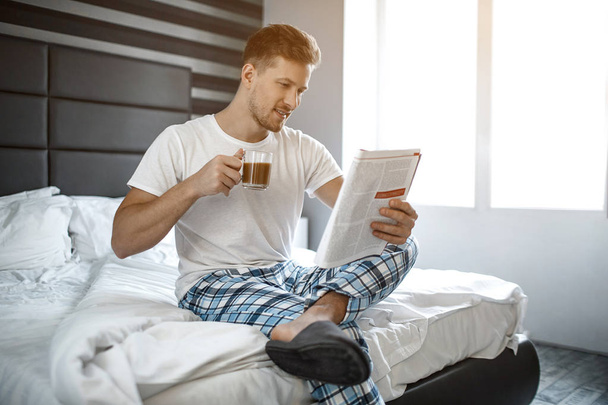 Young man on bed in the morning. He sit and read journal Guy hold cup of coffee. Nice and cheerful. - Foto, Bild