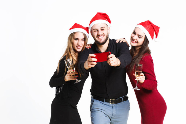 Two beautiful women and man in red christmas hats making selfie isolated on white background - Foto, imagen