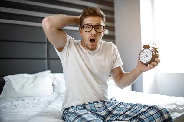 Amazed and scarec young man on bed in morning. He look emotionally on camera. Guy hold clock in hand. - Valokuva, kuva