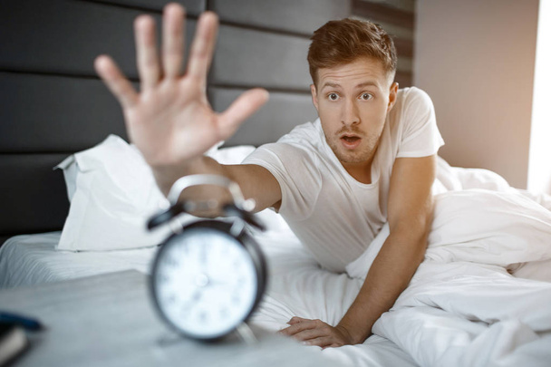 Young amazed man lyin on bed in morning. He overslept. Guy reach clock. - Photo, Image