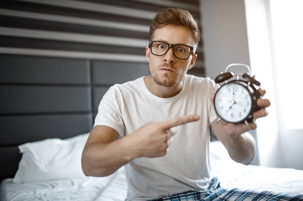 Serious angry young man on bed in morning. He look on camera and point on clock. - Zdjęcie, obraz