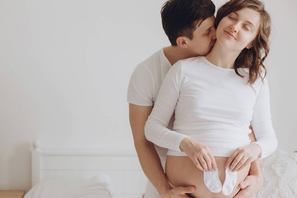 Happy young pregnant couple holding white little socks on belly  - Fotó, kép