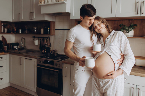 Happy young pregnant couple in white pajamas holding belly bump  - Φωτογραφία, εικόνα