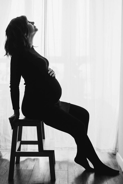 Silhouet van gelukkig zwangere vrouw in stijlvolle zwarte jurk sittin - Foto, afbeelding