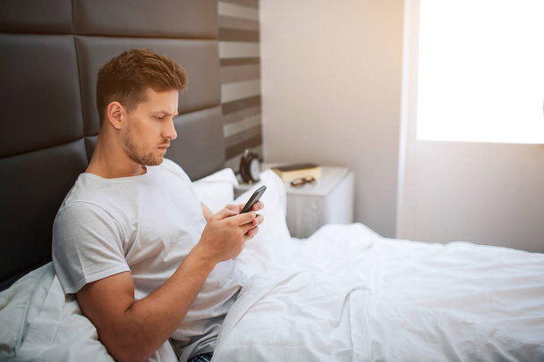 Young man in bed this morning. Serious thoughtful guy sit and look at phone. He hold ot in hands. Daylight. Early morning. - Fotografie, Obrázek
