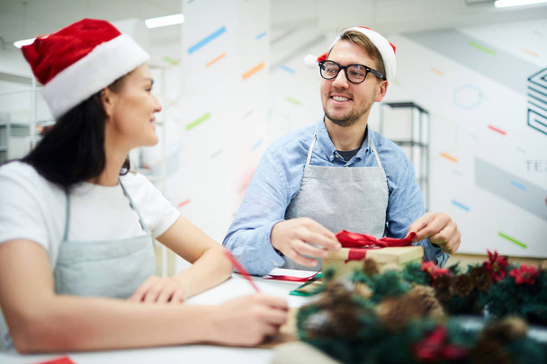 Šťastné a veselé mladé kolegy na klobouky a zástěrnách v Santa stolci sedí u stolu a povídají si o vánočních dárcích v kanceláři - Fotografie, Obrázek
