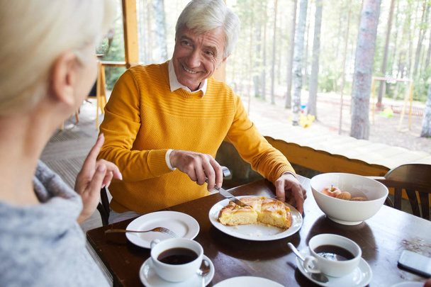 Szczęśliwy starszy człowiek cięcie jabłecznik podczas rozmowy z żoną przez herbatę w kawiarni - Zdjęcie, obraz