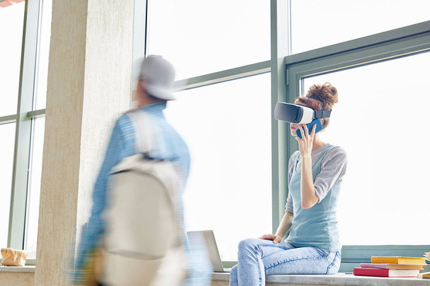 Menina estudante moderna em óculos VR sentado no peitoril da janela no corredor e falando no telefone celular no intervalo da universidade, movimento turvo do menino estudante com sacola
 - Foto, Imagem