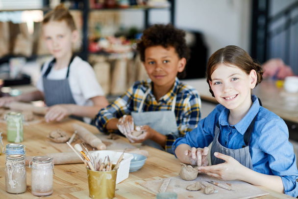 Menina feliz olhando para você enquanto sentado à mesa e fazendo xícara de barro com seus colegas de escola no fundo
 - Foto, Imagem