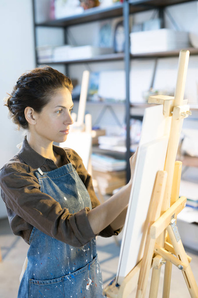 Молодая художница стоит перед мольбертом и рисует бумажный лист
 - Фото, изображение