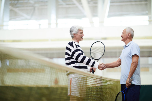 Two mature active tennis players with rackets handshaking over net after or before game on the court - Foto, Imagen