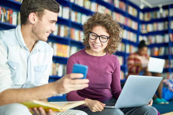 junger Mann zeigt seiner Klassenkameradin etwas auf seinem Smartphone, während sie Laptop in der Bibliothek benutzt - Foto, Bild