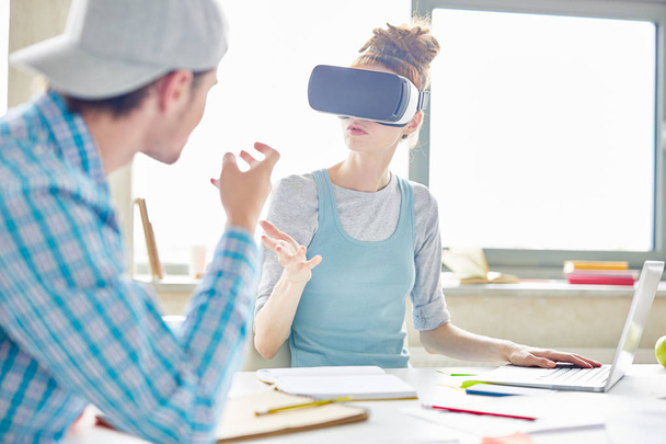 Compañeros de startup ocupados haciendo gestos de manos mientras discuten nuevo proyecto, mujer joven en gafas de realidad virtual explicando la estrategia a su colega
 - Foto, Imagen