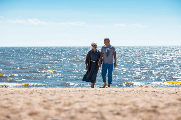 Ηλικίας ρομαντικό ζευγάρι έρχεται έξω από το νερό σε αμμώδη παραλία απολαμβάνοντας καλοκαιρινές διακοπές παραλία - Φωτογραφία, εικόνα