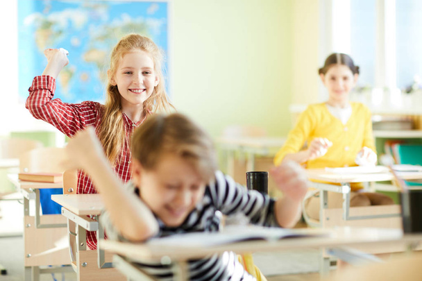 Crianças da escola animadas positivas jogando bolas de papel enquanto estavam sentadas em mesas e brincando em sala de aula
 - Foto, Imagem