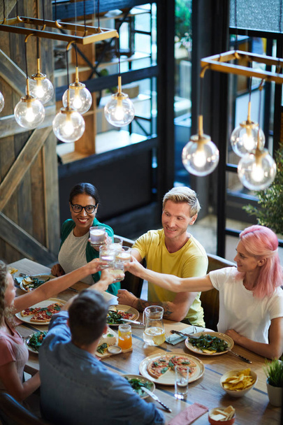 Grupo de jóvenes amigos felices brindando con bebidas sobre la mesa servida durante la cena en la cafetería
 - Foto, Imagen