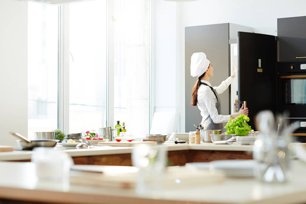 Visão traseira do chef feminino ocupado no chapéu e no avental, abrindo a geladeira e tirando os ingredientes enquanto se prepara para cozinhar na cozinha
 - Foto, Imagem