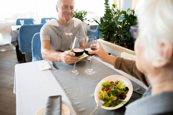 Over shoulder view of modern senior couple clinking wine glasses across table while enjoying date in cafe - Photo, Image