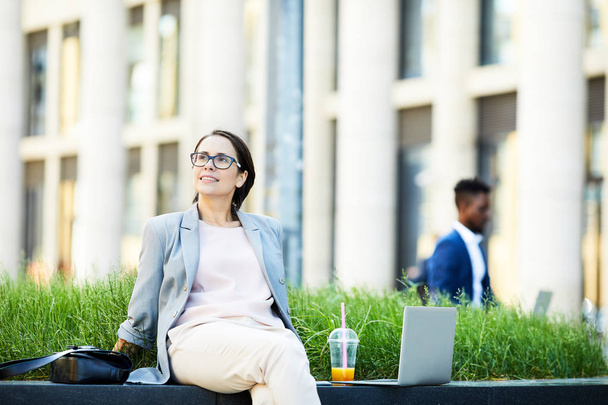 positiv entspannte junge Dame in Brille, die sich im Freien ausruht und auf Bank lehnt, während sie das Wetter genießt und sich in der Pause inspirieren lässt - Foto, Bild