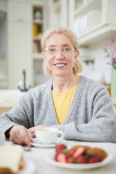 Heureuse femme blonde mature aux lunettes assise dans la cuisine, vous regardant et prenant le thé
 - Photo, image