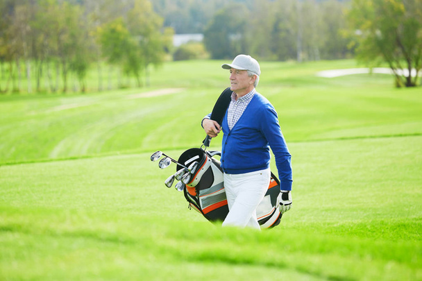 Homem sênior em casualwear andando para o jogo de golfe e carregando clubes no saco
 - Foto, Imagem