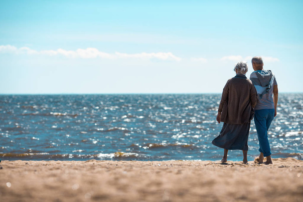 Вид ззаду літньої пари, що стоїть на піщаному пляжі і дивиться на море в сонячний день
 - Фото, зображення
