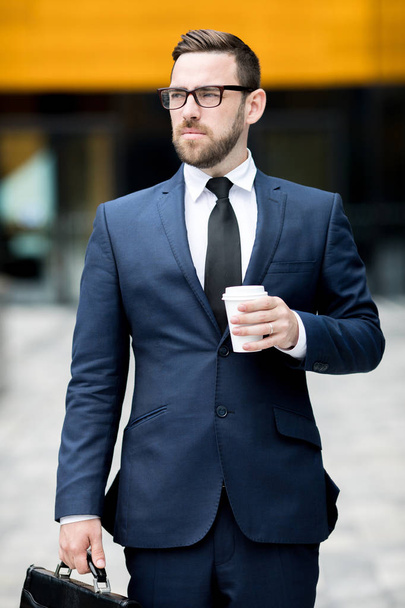 Молодой успешный красивый мужчина в деловом платье, очки держа чашку с горячим напитком и глядя в сторону
 - Фото, изображение