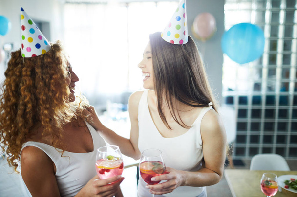Dos chicas amistosas alegres con bebidas riendo mientras se divierten en la fiesta de cumpleaños
 - Foto, Imagen