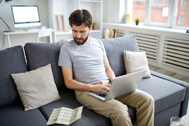 Jovem contador olhando para os dados financeiros em papel wnd inseri-lo no laptop enquanto trabalhava no sofá no home office
 - Foto, Imagem