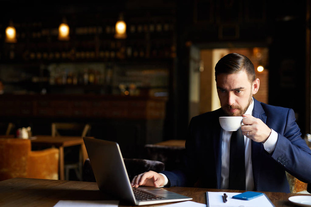 Serieuze doordachte knappe bebaarde zakenman in pak werken aan verslag en koffie drinken terwijl zittend in lege restaurant - Foto, afbeelding