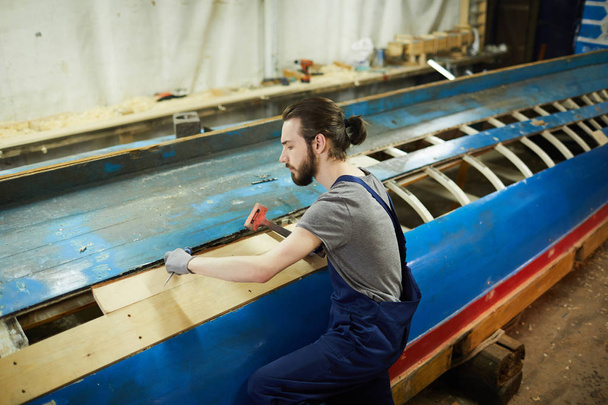 Trabajador joven sentado junto a un gran recipiente sin terminar y martilleando tablas de madera
 - Foto, Imagen