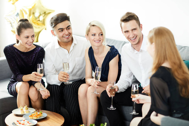 Группа веселых друзей в умной одежде, сидящих на диване, пьющих шампанское и разговаривающих на вечеринке
 - Фото, изображение