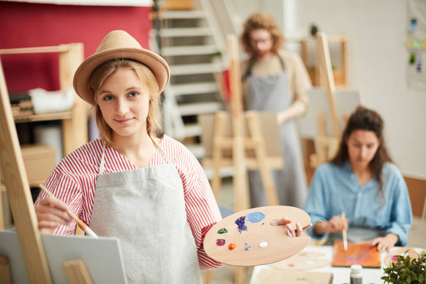 Young creative woman with palette and paintbrush painting in studio of arts - Photo, Image