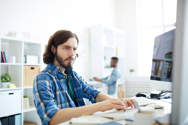 Серьезный концентрированный хипстер молодой бородатый программист, работающий с двумя настольными компьютерами и печатающий на клавиатуре, сидя за столом в офисе
 - Фото, изображение